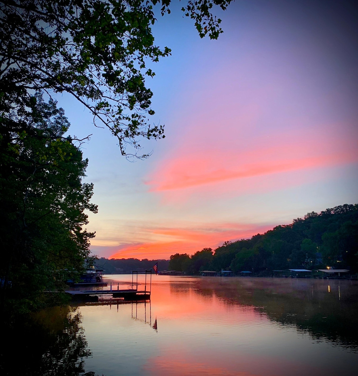 A serene lake view is framed by lush trees, with soft pink and purple hues reflecting on the water's surface. The calming scene is enhanced by docked boats on the shore, and a gentle mist rises from the lake during twilight.