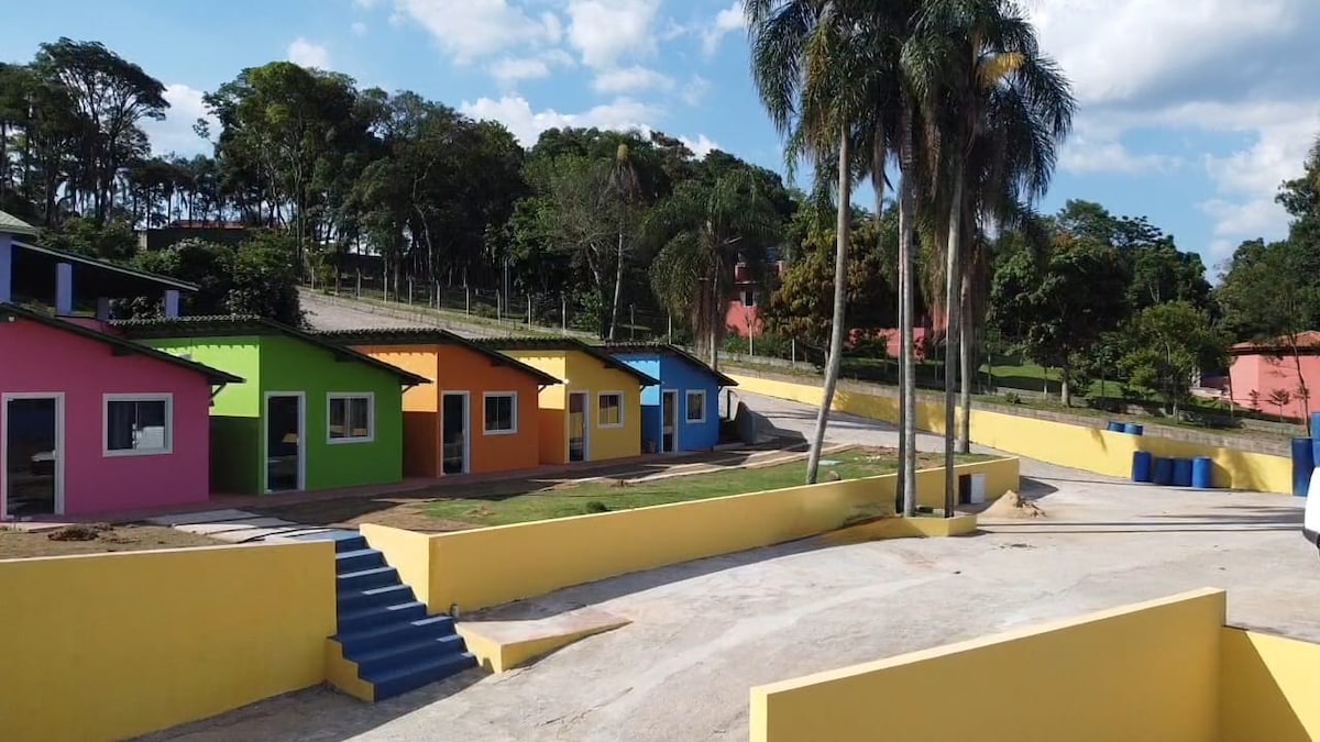 A colorful cluster of charming cottages is set against a backdrop of greenery. Each cottage features a distinct vibrant color, with large windows allowing natural light. The area is bordered by well-maintained landscaping, creating an inviting and cheerful environment.