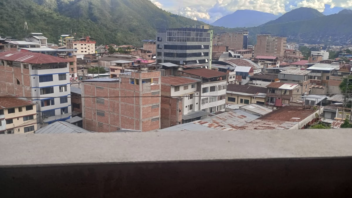 A view from a balcony reveals a mix of brick and concrete buildings nestled against a backdrop of lush mountains. A varied skyline can be seen, featuring rooftops of different materials and colors, with natural greenery complementing the urban setting.