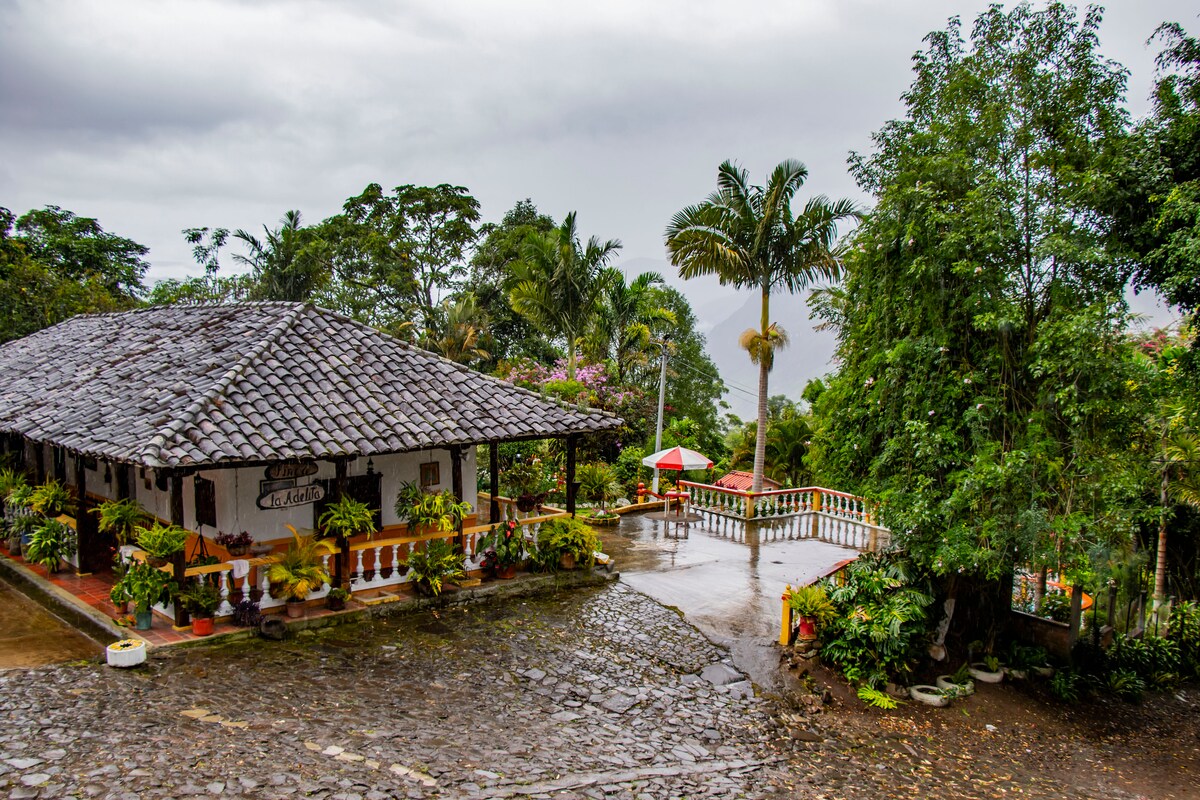 A charming building with a traditional tiled roof is set amidst lush greenery. The stone pathway leads to a spacious terrace adorned with vibrant plants and seating areas, offering a peaceful view of the surrounding mountains under a cloudy sky.