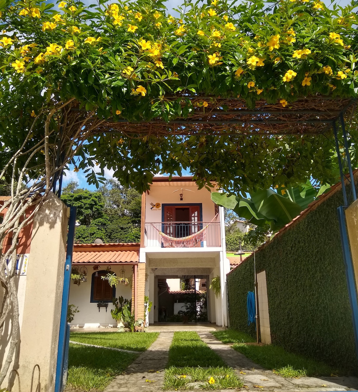 A welcoming entrance is framed by vibrant yellow flowers, leading to a two-story house. Green foliage and a stone pathway create a serene ambiance. The house displays a balcony with decorative accents, and ample natural light brightens the entrance adorned with wooden doors.