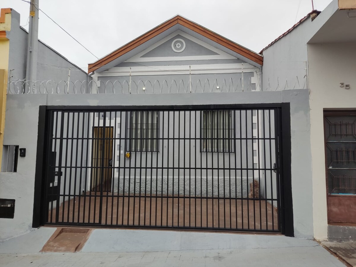The exterior of the kitinete features a classic-style facade with a distinctive roof design. A secure black metal gate is visible, enclosing a paved area in front of the property. Two windows are adorned with grilles, enhancing security while allowing natural light.