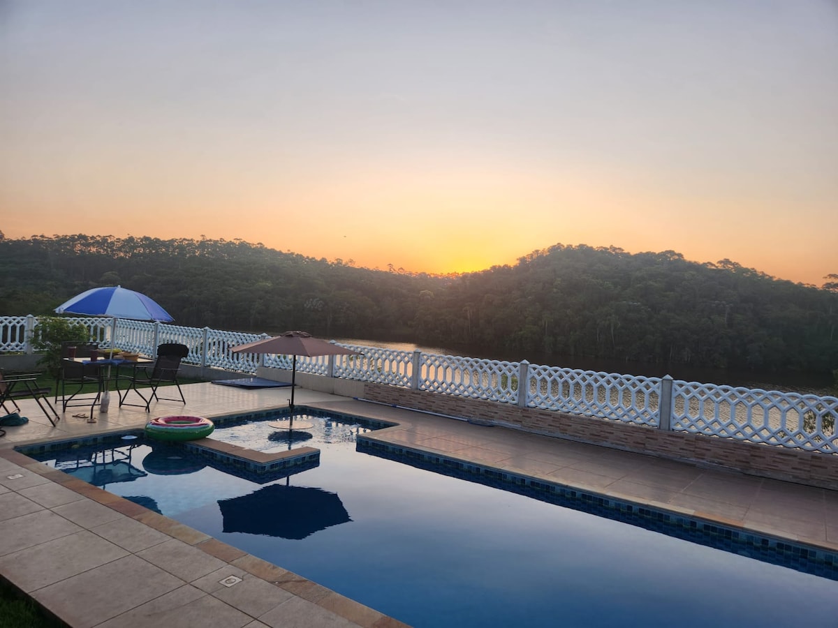 An outdoor area showcases a large swimming pool with an integrated jacuzzi, framed by a serene landscape. A patio featuring umbrellas and chairs overlooks a peaceful reservoir, with trees lining the horizon under a soft sunset glow.