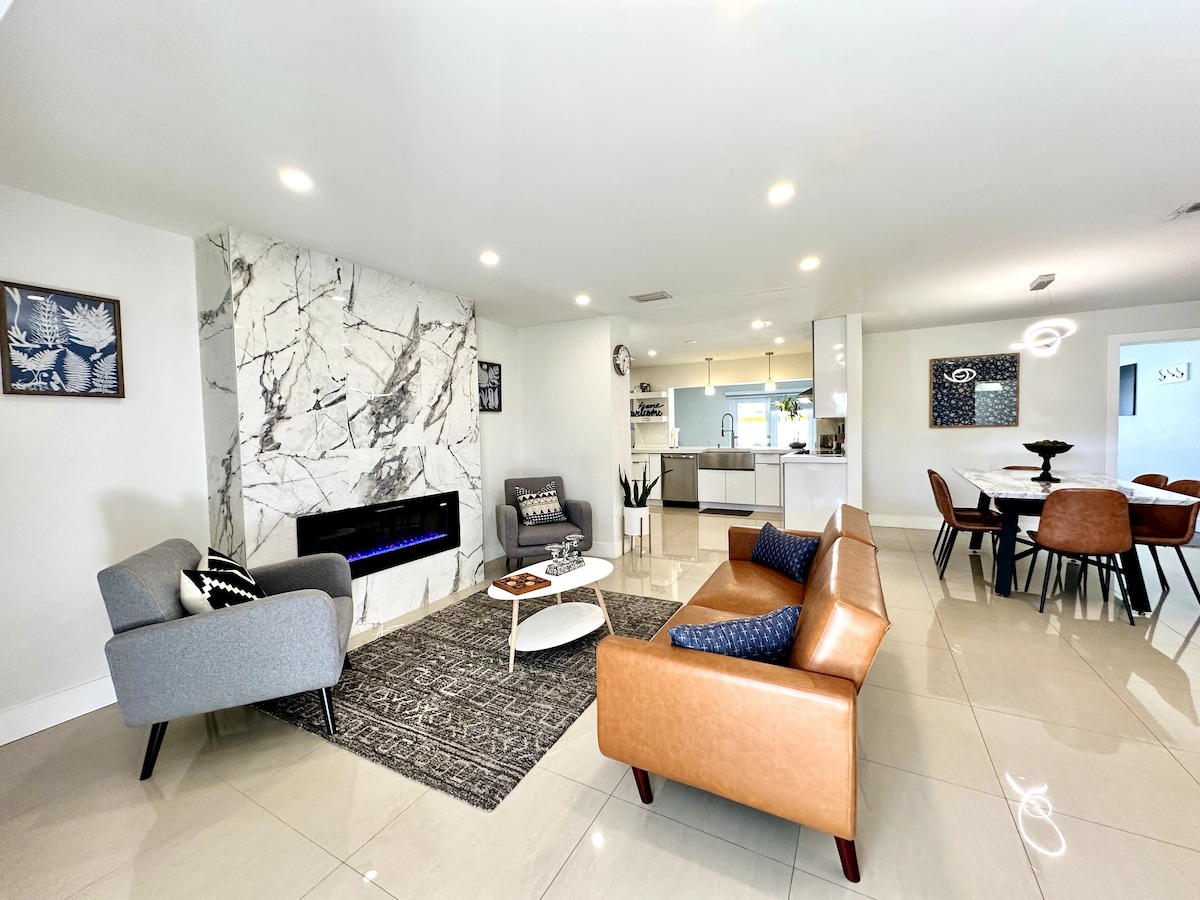 A modern living area features a mix of comfortable seating options, including a light grey chair and a brown couch. A sleek marble accent wall serves as a backdrop for a linear electric fireplace. A round dining table with stylish chairs is visible in the background.