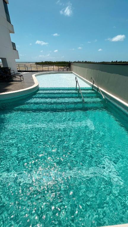 A swimming pool extends alongside the building, featuring gentle steps leading into the clear, turquoise water. Sunlight reflects off the surface, creating a shimmering effect. The pool area offers views of the surrounding landscape, with a hint of the ocean visible in the distance.