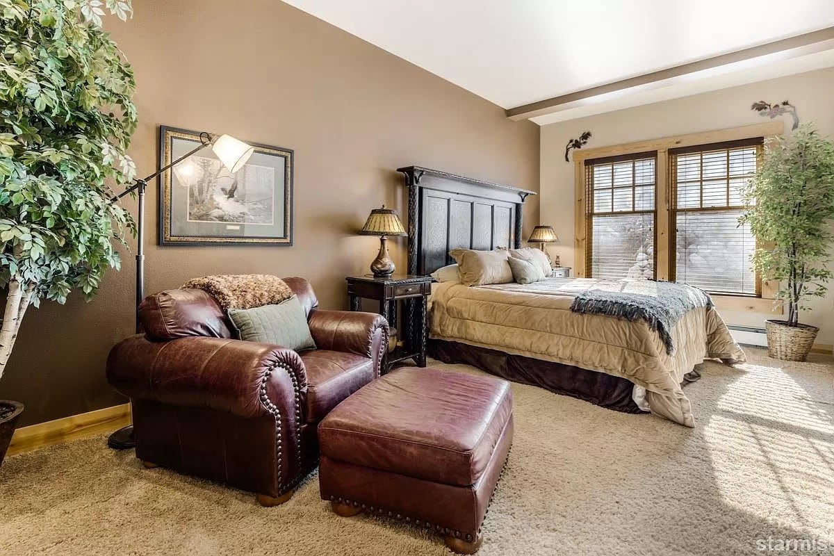A spacious master bedroom features a king-size bed with a decorative quilt and plush pillows. A leather armchair and ottoman provide cozy seating. Natural light filters through two windows, flanked by artificial plants, enhancing the warm atmosphere.