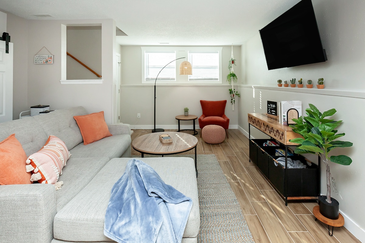 A spacious living area features a comfortable sectional sofa with decorative orange pillows. A coffee table sits at the center, complemented by a cozy accent chair and a potted plant. Natural light enters through the windows, enhancing the inviting atmosphere of the space.