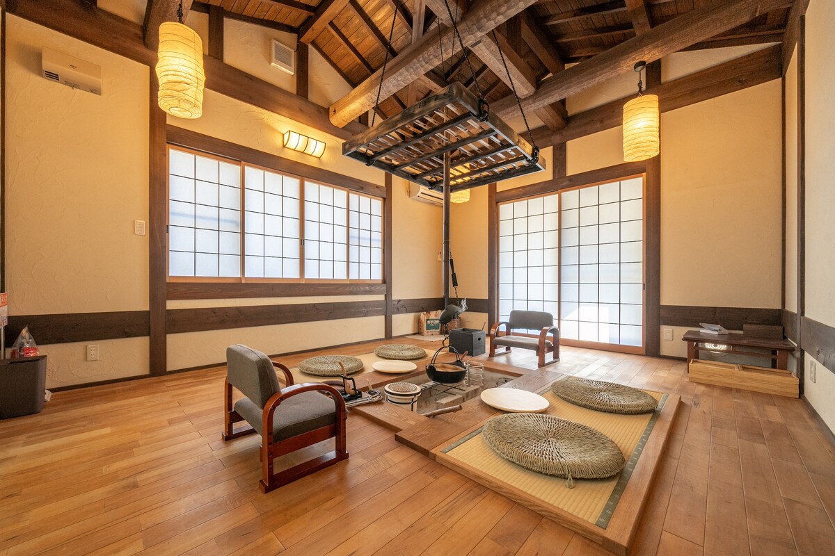 The spacious room is adorned with wooden beams and a high ceiling. Traditional tatami mats are arranged alongside low tables and cushions, fostering a cozy seating area. Large shoji windows allow natural light to filter in, enhancing the serene atmosphere.