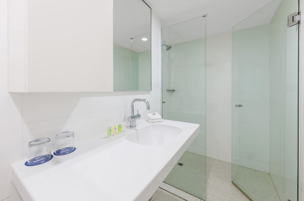 A modern bathroom features a spacious sink counter with neatly arranged toiletries. A glass shower enclosure is visible, along with white tiled walls that enhance the bright and clean appearance. Ample lighting illuminates the area, creating a fresh and airy ambiance.