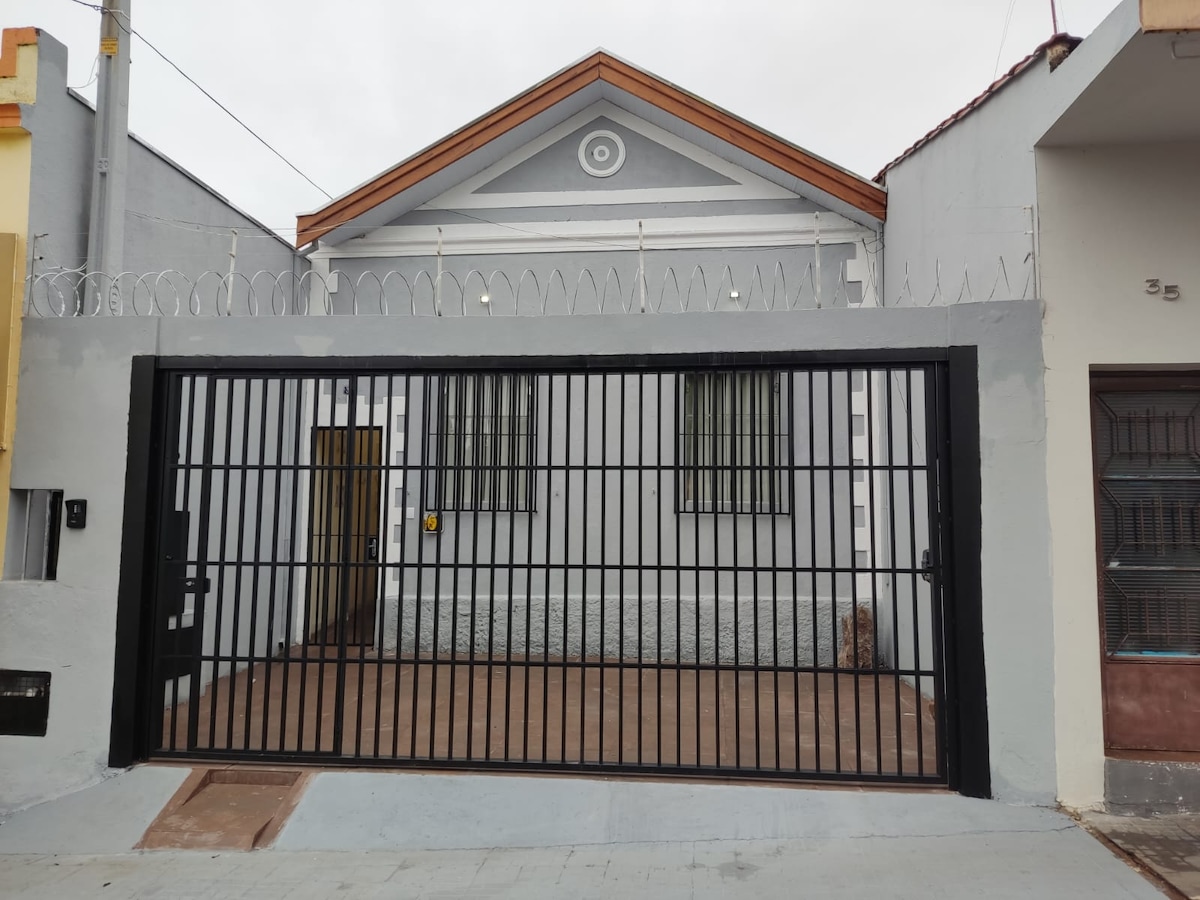 An exterior view of the property is presented, featuring a traditional house facade with a distinctive architectural detail. A secured metal gate is visible, providing access to the enclosed courtyard area, which is set against a solid wall. The overall setting is quiet and inviting.