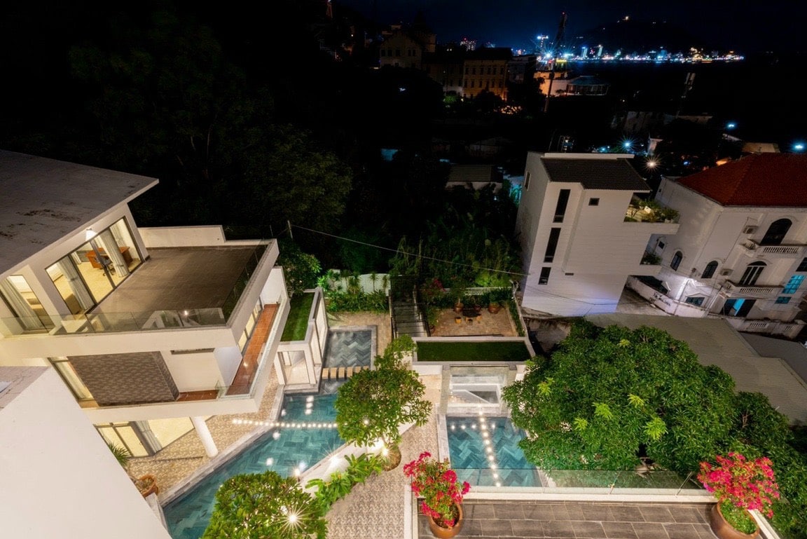 A night view reveals a modern villa with multiple levels, accented by soft lighting. A paved pathway leads to the inviting pool area, surrounded by lush greenery and vibrant flowers. The city skyline glimmers in the background, creating a serene atmosphere.