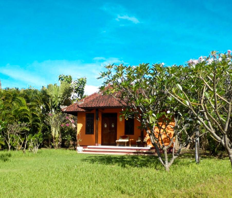 A charming bungalow is nestled within a lush green garden, surrounded by vibrant tropical foliage. The structure features a traditional roof and a welcoming entrance, framed by flowering plants and a spacious lawn that enhances the serene setting.