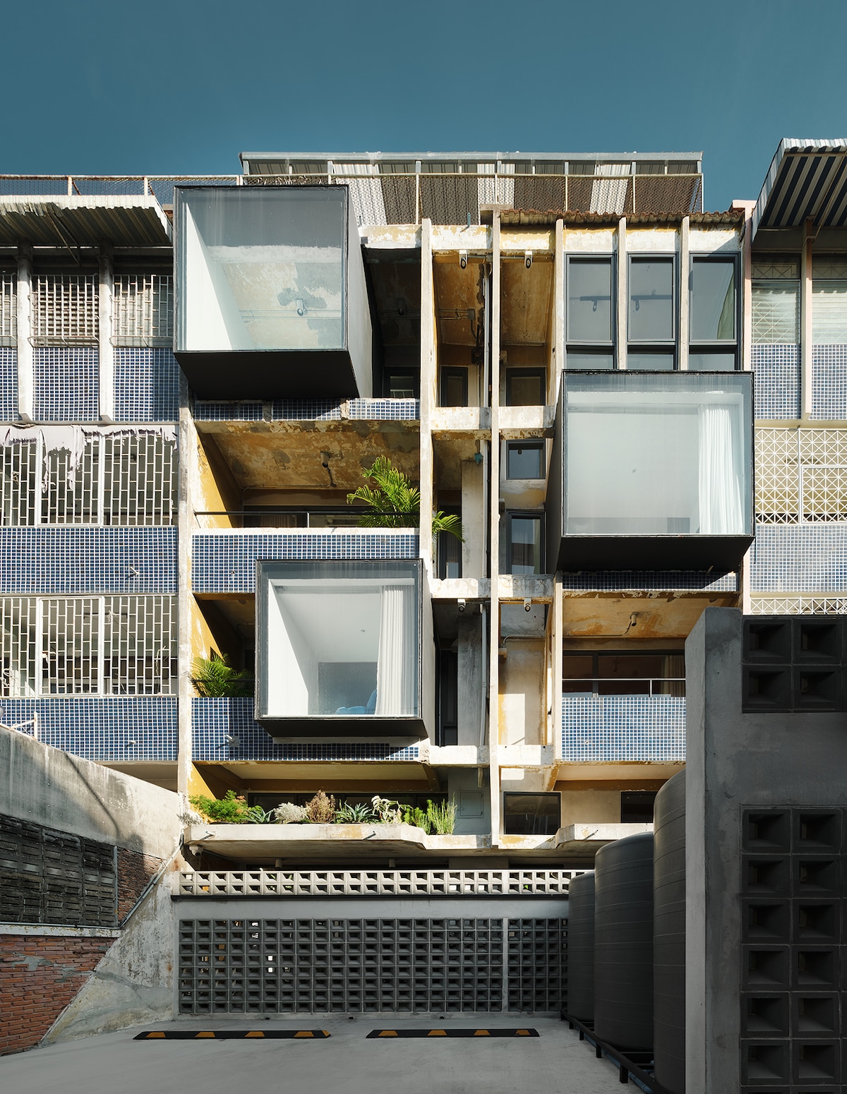 The exterior of a modern building showcases a unique architectural design with cantilevered glass cubes. Exposed concrete and vibrant patterned tiles create a striking contrast, while greenery is integrated into the structure. The upper levels feature large windows that allow natural light to illuminate the interior spaces.