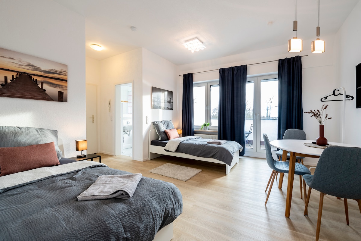 A modern studio layout features two spacious beds with neutral bedding. Natural light fills the room through large glass doors, appealingly framed by dark curtains. A round dining table, accompanied by blue chairs, offers a comfortable space for meals or work.