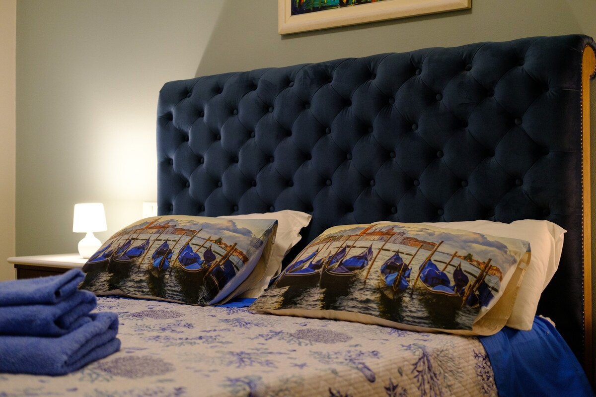 A spacious bed with a tufted blue headboard is presented against a soft green wall. Decorative pillows feature a nautical design, complemented by a light blanket atop the bed. A bedside lamp emits a gentle glow, and neatly arranged towels are accentuated in the foreground.