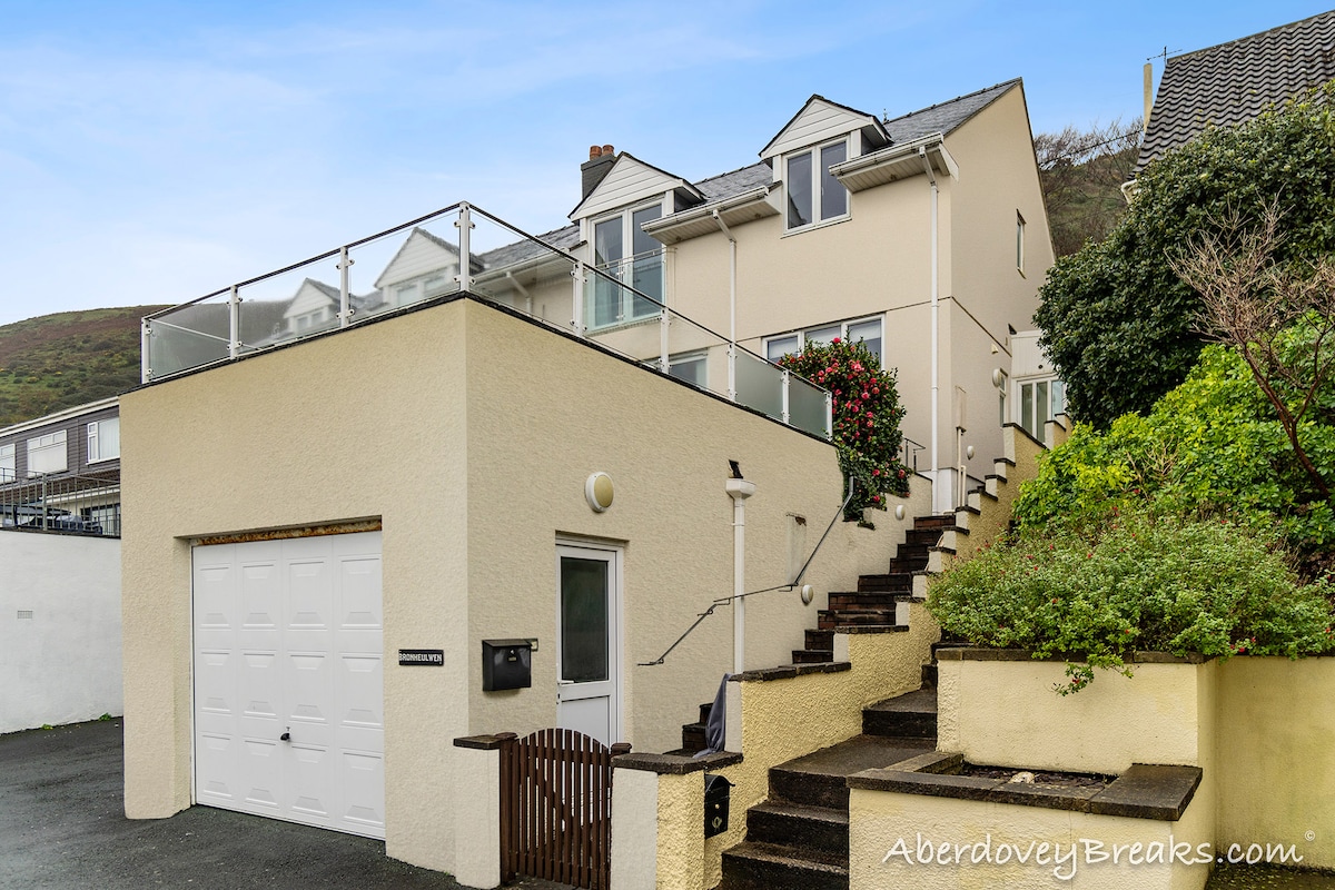 The exterior of the coastal property is presented, showcasing a light-colored building with multiple windows. A private driveway is visible, leading to a garage. Stairs ascend from the parking area to a top terrace, surrounded by greenery.