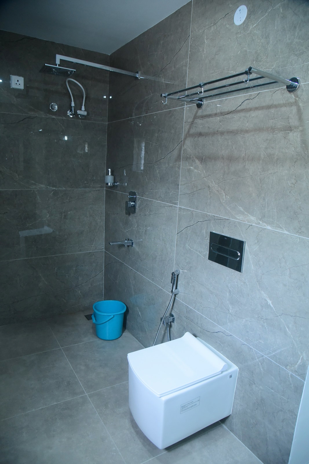 A modern bathroom is showcased with sleek gray tiles covering the walls and floor. A compact toilet is positioned beside a shower area equipped with multiple showerheads. A blue bucket sits in the corner, adding an element of functionality.