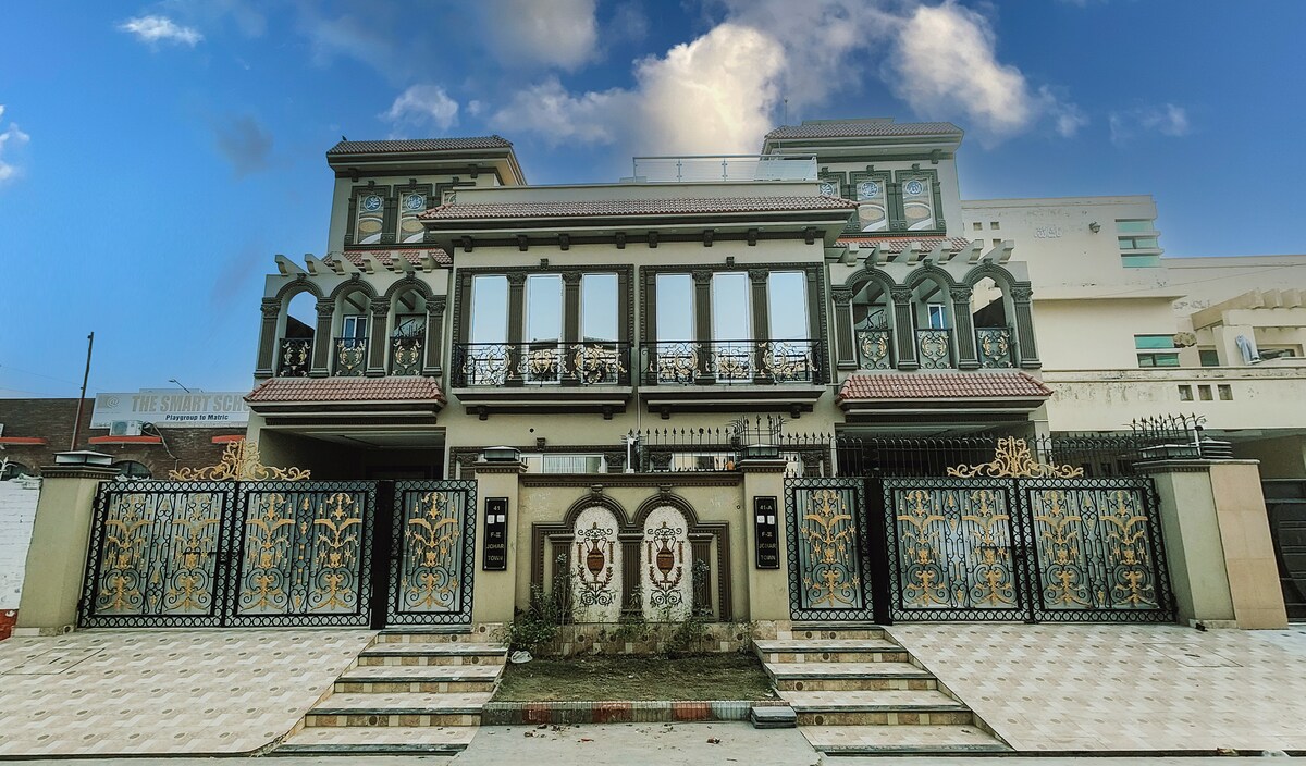 The exterior of the property showcases a two-story structure with intricate architectural details and a decorative facade. Large windows reflect the surrounding sky, while ornate gates provide security and an inviting entrance, framed by paved walkways leading to the main entrance.