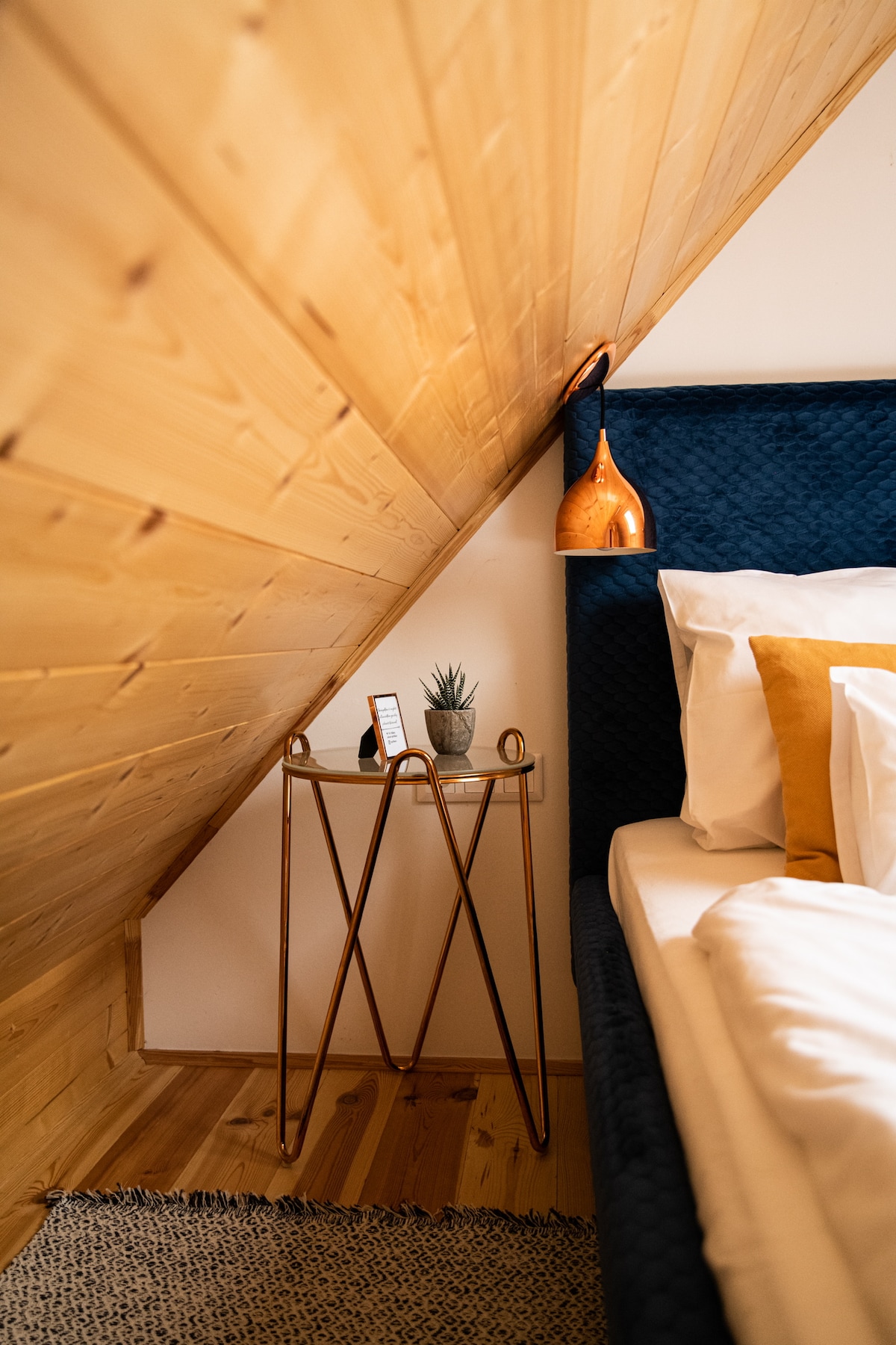 A cozy bedroom corner features a low angled ceiling with wooden paneling. A plush dark blue bed is complemented by bright pillows. A small metallic side table holds a decorative plant and a minimalist clock, adding a touch of modern simplicity.