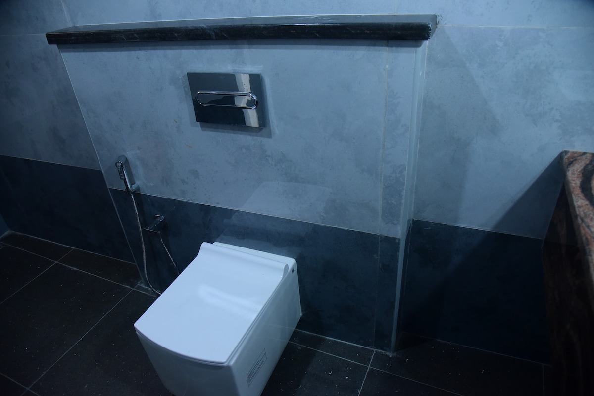 A modern bathroom features a sleek, wall-mounted toilet with a dual flush mechanism. The walls are finished in a combination of soft gray tones, complemented by dark marble accents. A stylish sink with a polished countertop can be seen in the corner, enhancing the contemporary design.