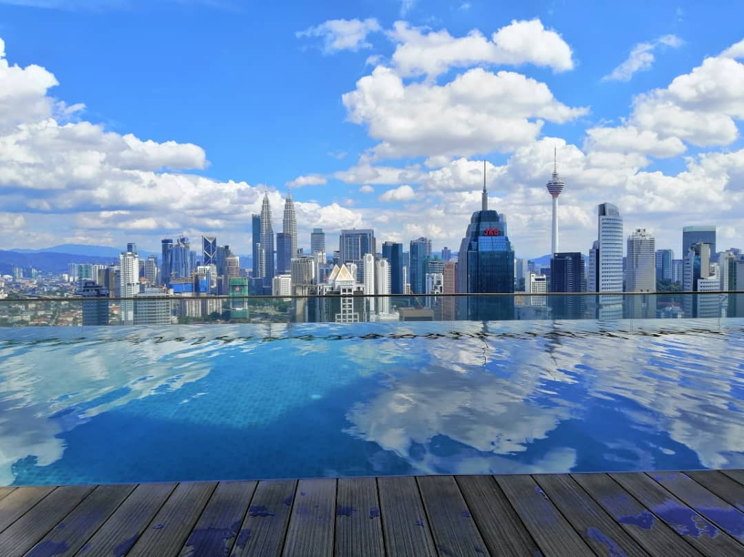 An infinity swimming pool is visible in the foreground, reflecting the skyline of Kuala Lumpur. The backdrop features a clear blue sky with scattered clouds and modern city buildings, including notable towers, creating a scenic urban landscape.