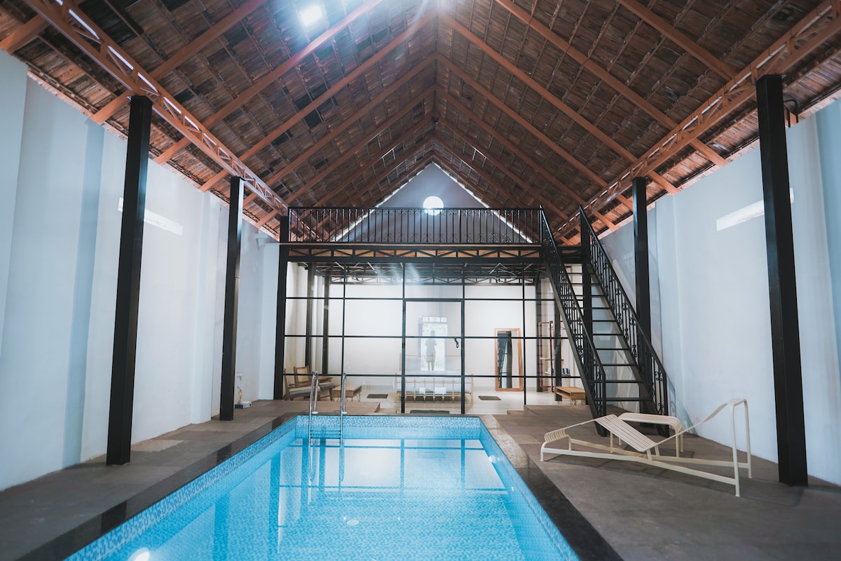 An indoor area featuring a private pool with clear blue water. A spacious high ceiling showcases exposed wooden beams. A modern metal staircase leads to an upper level, while lounge chairs are positioned beside the pool, offering space for relaxation.