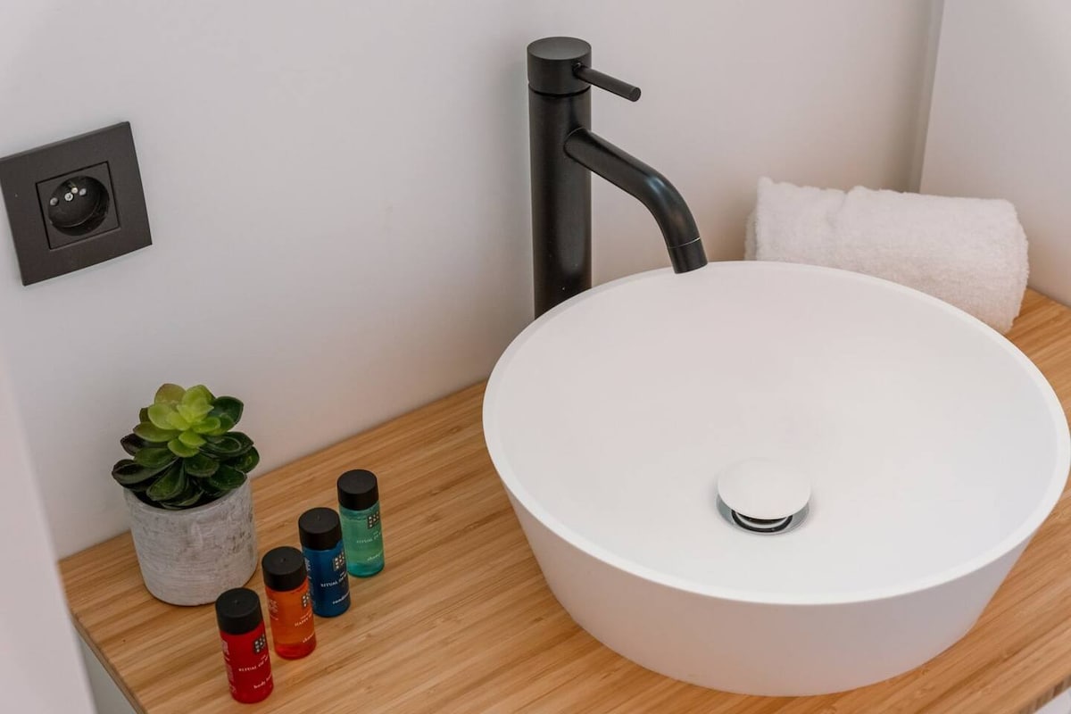 A modern bathroom sink is elegantly positioned on a bamboo countertop. A round white basin is paired with a sleek black faucet. Beside the sink, a small plant adds a touch of greenery. Several colorful toiletries are neatly arranged nearby, along with a white towel.