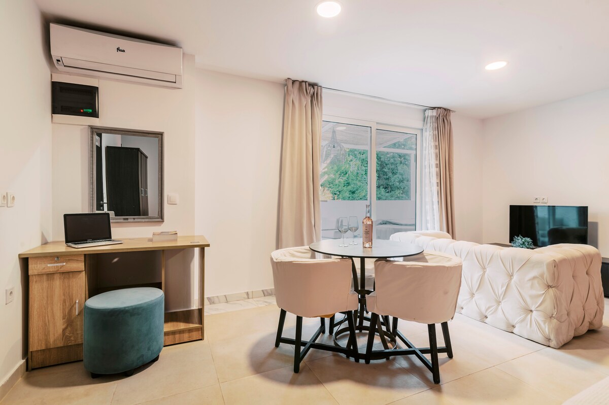A modern living area is furnished with a tufted white couch and a glass dining table surrounded by four chairs. A desk with a chair is positioned beside a large window, and a television is mounted on the wall opposite the seating arrangement.