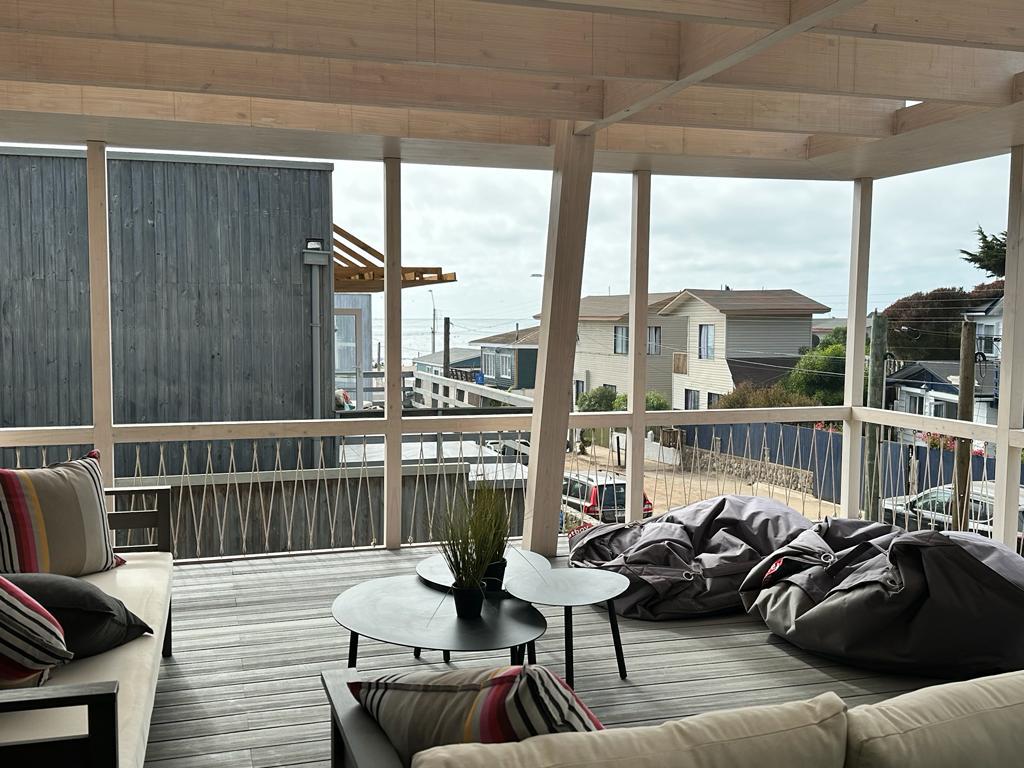 The terrace features a wooden deck with expansive views of the surroundings. Comfortable seating is arranged around a central table, accompanied by bean bags. Natural light floods the area through large openings, creating a spacious and inviting atmosphere.