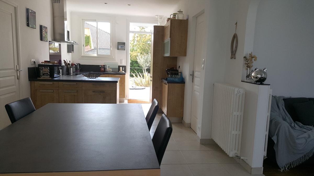 The kitchen features wooden cabinetry and a spacious dining area with a dark table and black chairs. Large windows allow natural light to brighten the space. A cozy seating area is visible, adorned with a throw blanket and decorative elements.