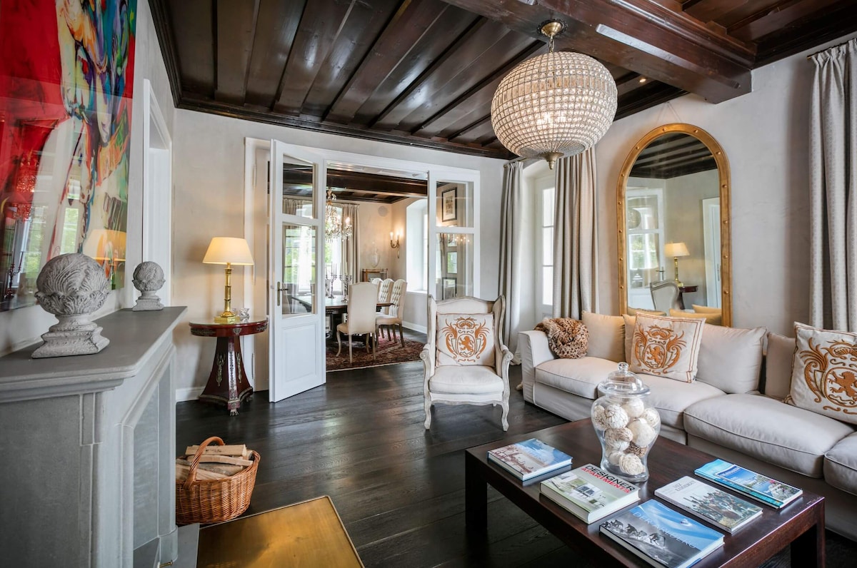 The living room features a cozy seating area with a light-colored sofa adorned with decorative cushions. A large mirror and elegant chandelier enhance the room's charm. Double doors lead to a dining area, while natural light filters through the curtains, highlighting the wooden ceiling.