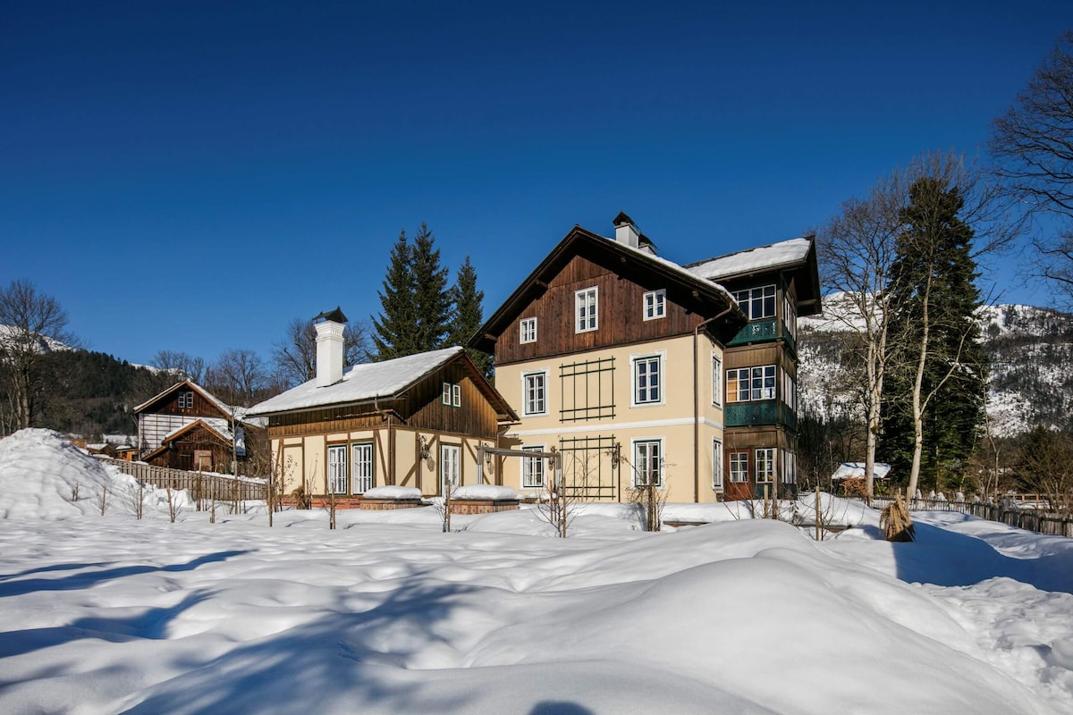 The exterior of a charming alpine villa is displayed, surrounded by a blanket of snow. The building's diverse architectural styles and colors stand out against the clear blue sky, with tall trees framing the view in the background.