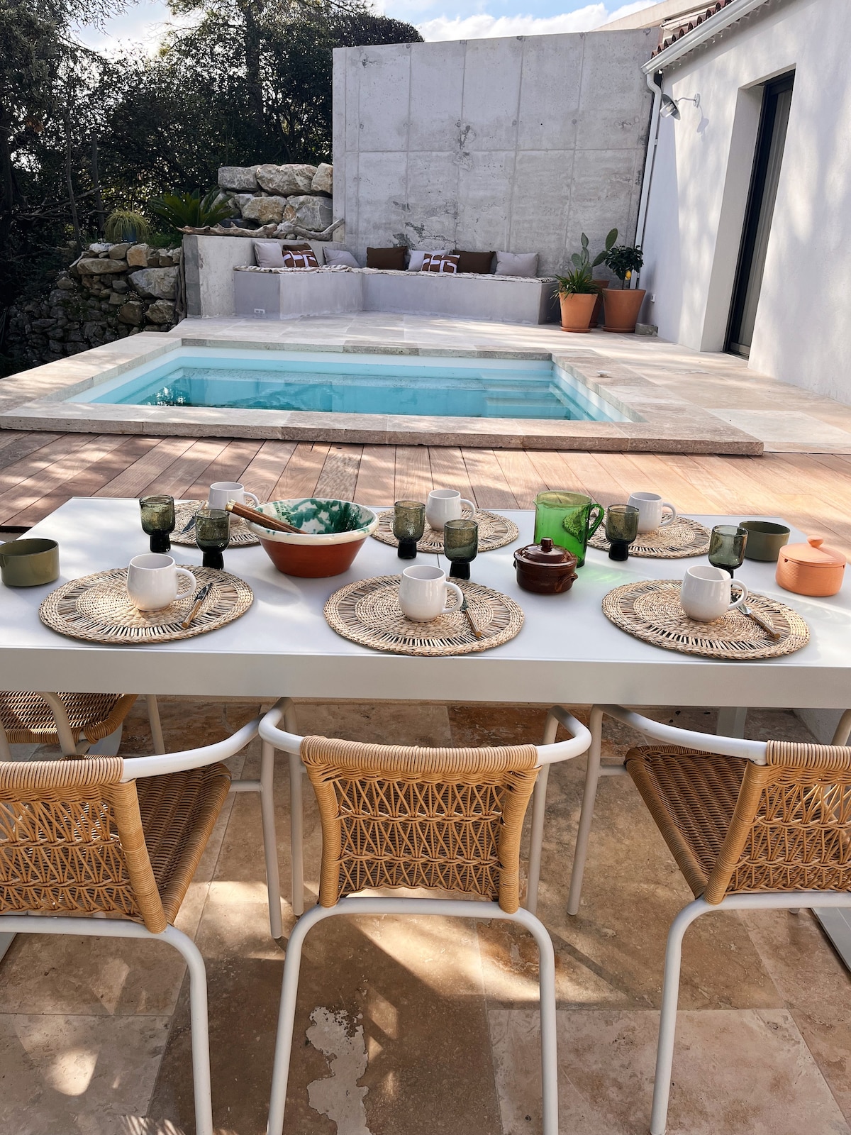 A dining table set for a meal features woven placemats and ceramic dinnerware, complemented by various glassware in shades of green. In the background, a small pool is surrounded by a wood deck and planters, offering a serene outdoor setting.
