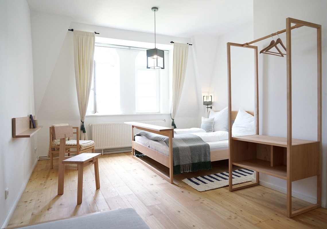 A bright room is featured with large windows allowing natural light. The space includes a spacious bed draped with a soft blanket and a bedside lamp. A wooden wardrobe and a small seating area with a chair are positioned adjacent to the bed.