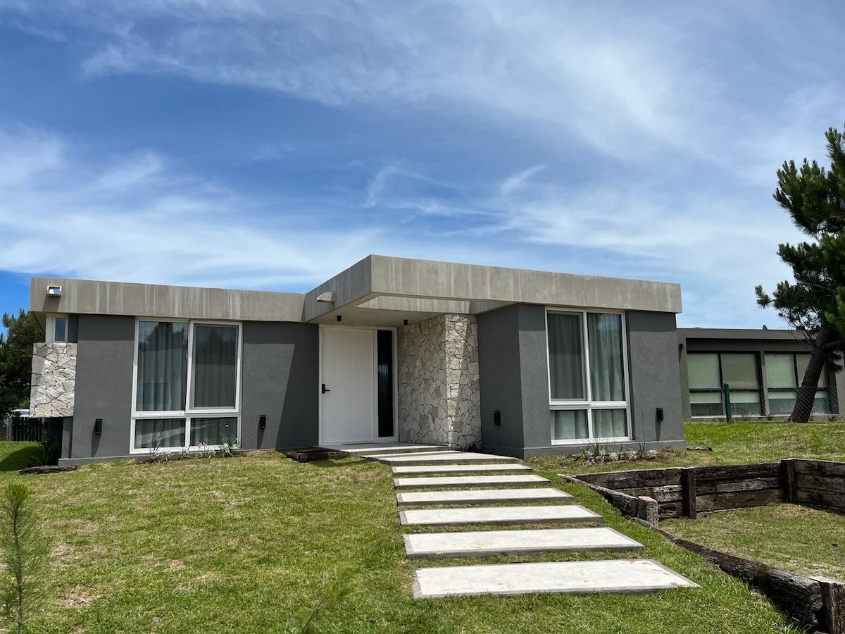 A modern one-story house is presented with a smooth concrete facade and large glass windows, allowing natural light to fill the interior. A landscaped front yard features a well-maintained lawn and a stone pathway leading to the entrance.