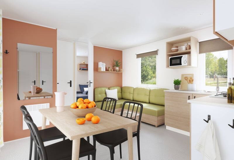 A cozy dining area is furnished with a wooden table surrounded by black chairs. A green seating area is adjacent, offering soft cushions and a view to outside. Light enters through large windows, and a microwave is visible on the shelf in the kitchen area.