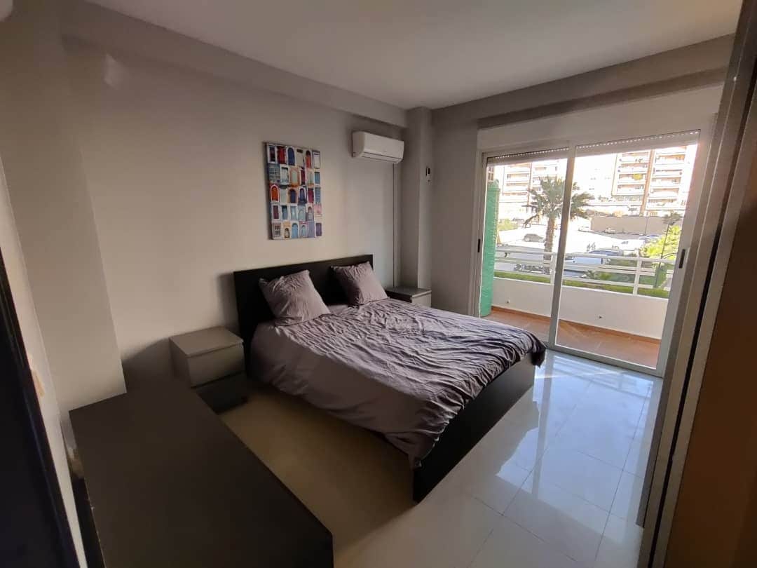 A spacious bedroom is outfitted with a large bed featuring a neutral-hued bedding set. Two nightstands are positioned beside the bed, and a colorful wall art piece adds visual interest. Large windows provide natural light and offer a view of the outside balcony.