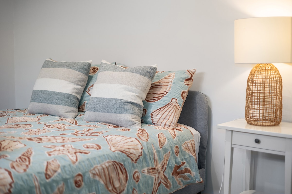 A cozy sleeping area features a bed adorned with a shell-patterned coverlet. Decorative pillows in varying textures enhance the bedding. A white nightstand accommodates a woven lamp, providing soft illumination against the neutral wall.