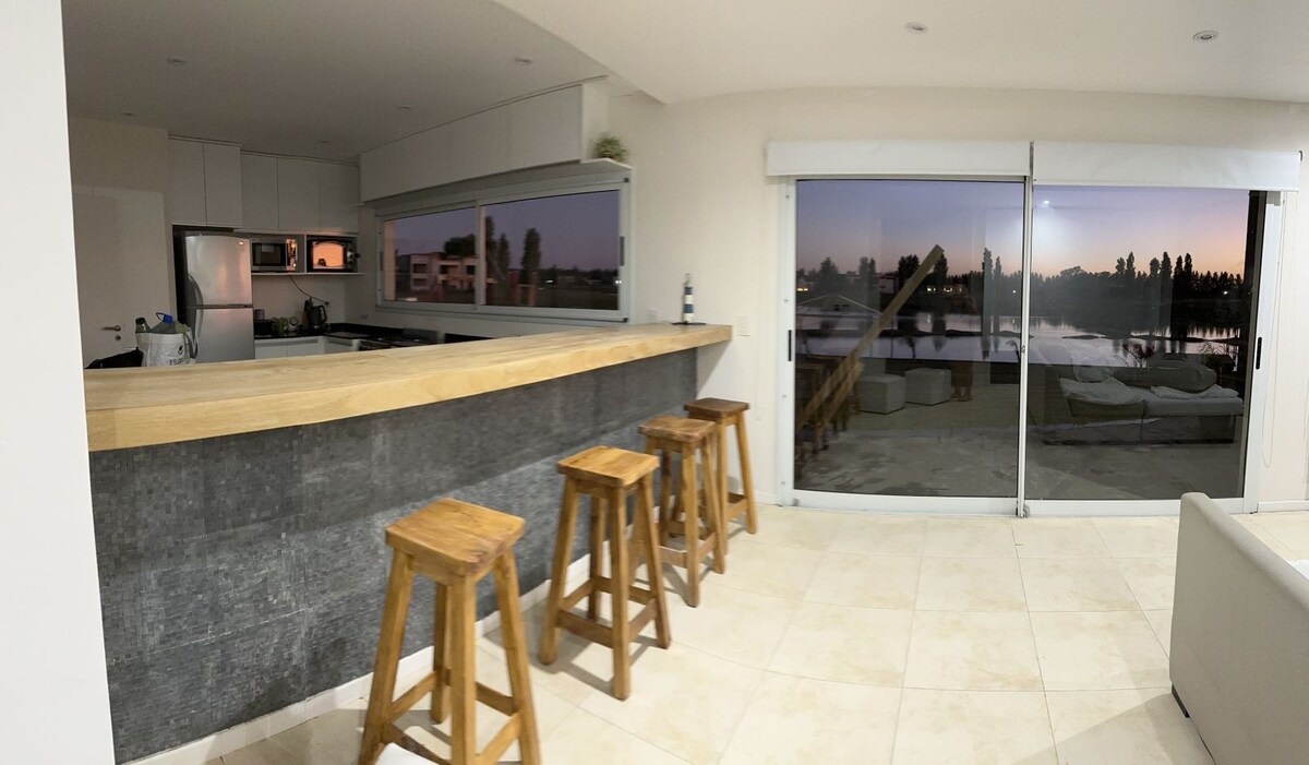 The open kitchen area features a wooden countertop with four high stools positioned alongside it. Large glass sliding doors provide a view of the tranquil lake outside, enhancing the bright and airy ambiance of the space.