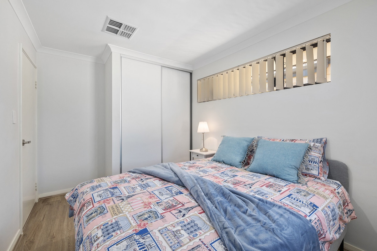 A sleeping area features a spacious bed adorned with a patterned quilt and accent pillows. Soft lighting emanates from a bedside lamp, while a sleek wardrobe is positioned against the wall. Light filters through a decorative window above, adding a touch of elegance.