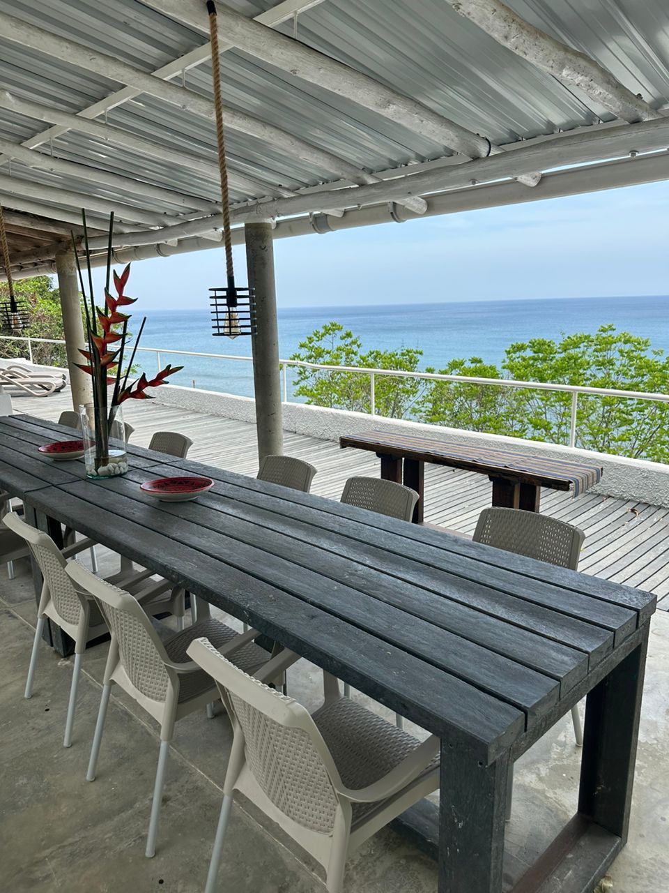An outdoor dining area is presented with a long wooden table and multiple light-colored chairs. A vase with tropical flowers adorns the table, while the ocean view stretches in the distance, with greenery lining the perimeter.