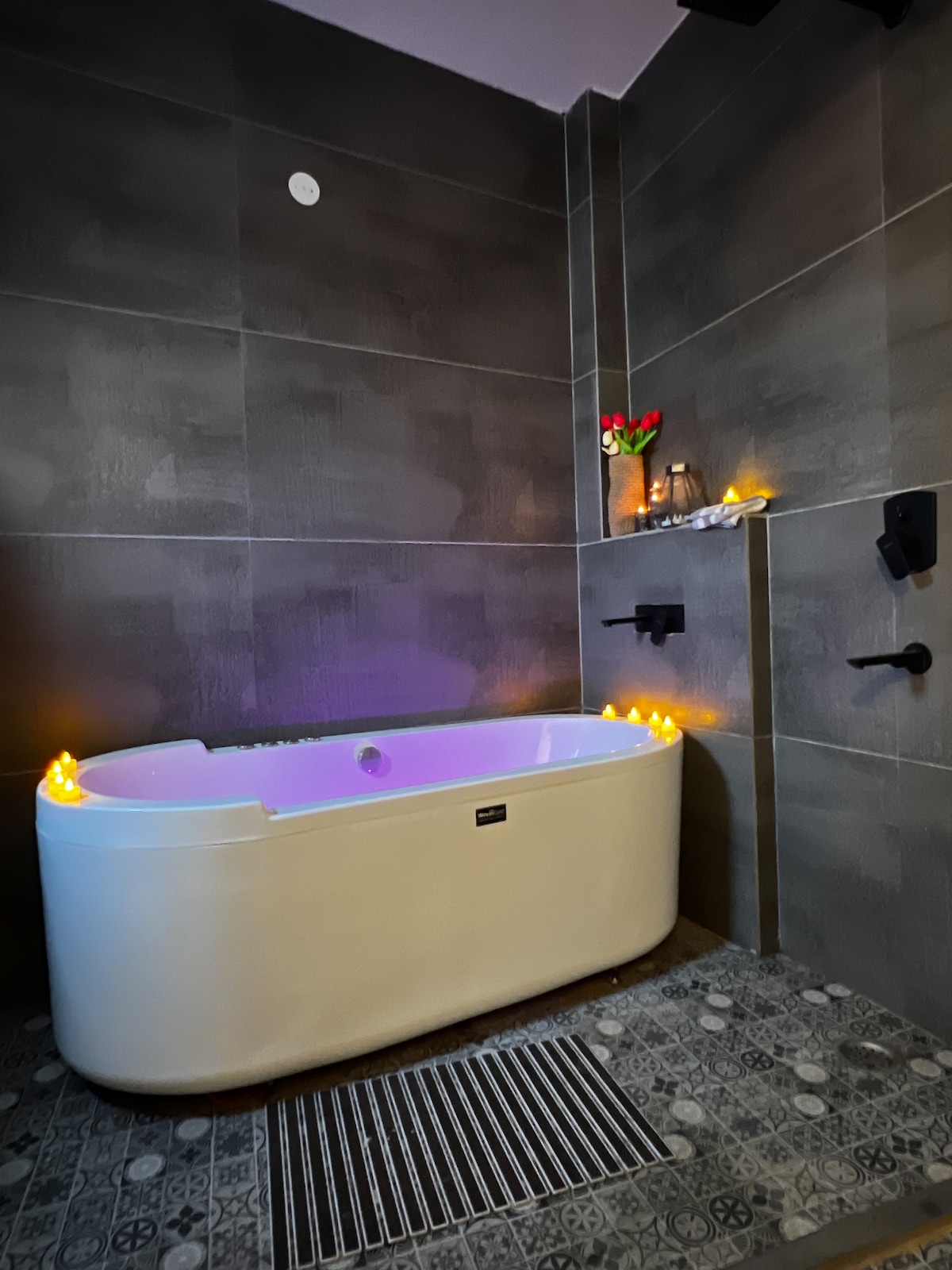 A modern bathtub is positioned against a gray tiled wall, adorned with candles that emit a gentle glow. Fresh flowers are placed nearby, and minimalistic fixtures complement the contemporary design of the space.