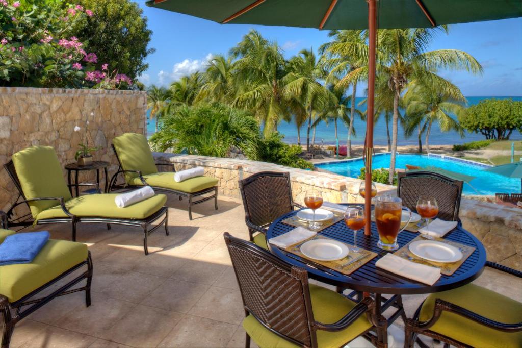 An outdoor dining area is set with a round table and ten chairs, complemented by green loungers under a large umbrella. A view of the ocean and palm trees is visible in the background, with a pool nearby, suggesting a relaxing environment.