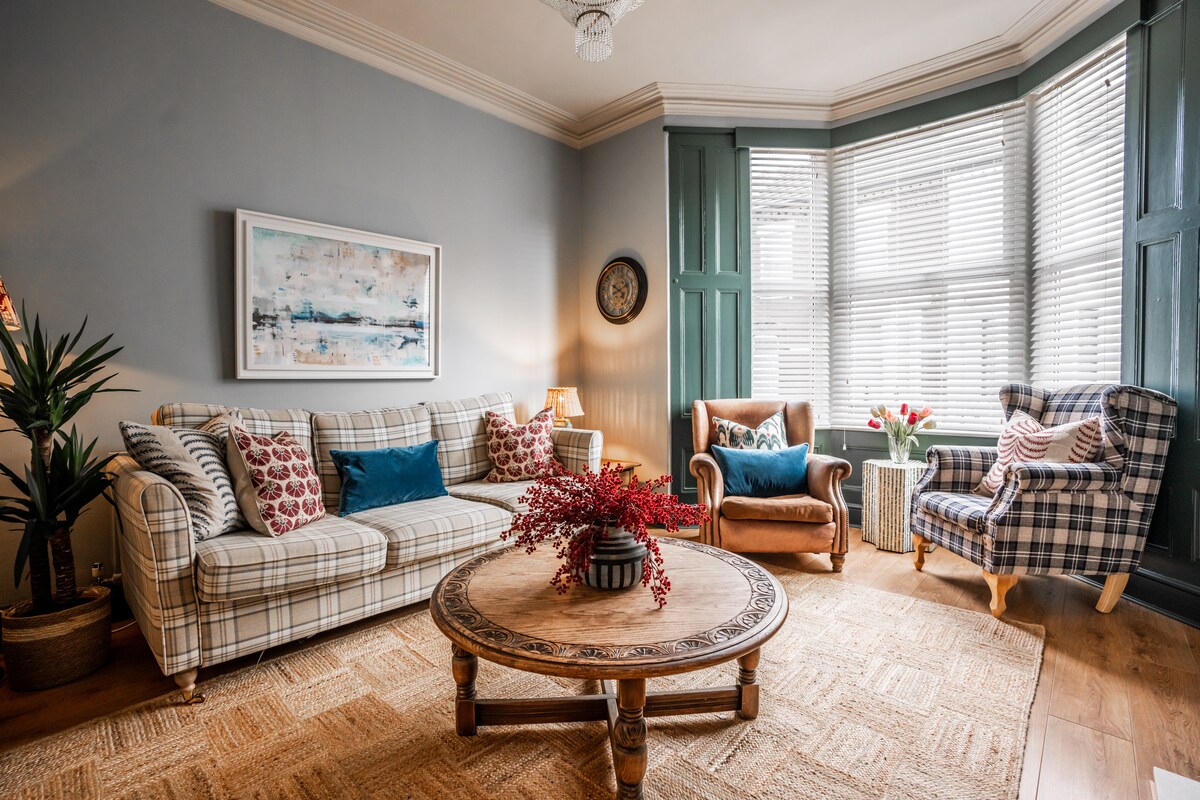 A welcoming living room features a large round wooden coffee table surrounded by a variety of comfortable seating options, including a patterned sofa and an upholstered armchair. Natural light streams in through bay windows adorned with blinds, enhancing the soft color palette of the space.