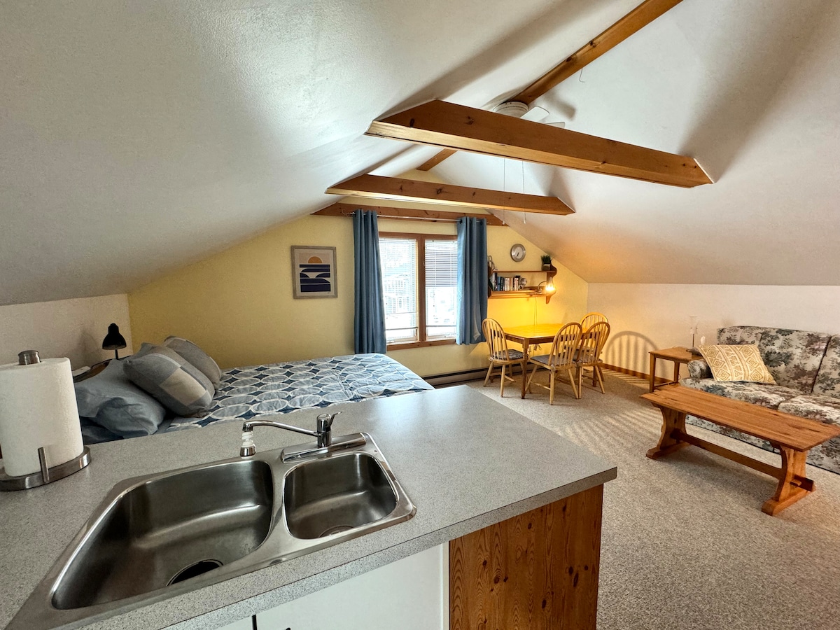 An open-concept loft showcases a spacious sleeping area with a cozy bed dressed in blue and white bedding. A dining table with four chairs and a living area with a patterned couch create a versatile space, illuminated by natural light from a window.