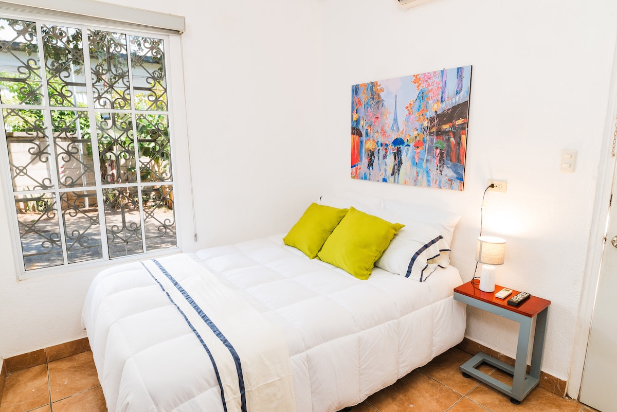 A bright bedroom features a spacious bed with white bedding and two green pillows. A colorful painting adorns the wall above the bed. Natural light enters through a window with decorative ironwork, enhancing the minimalist decor and peaceful atmosphere.