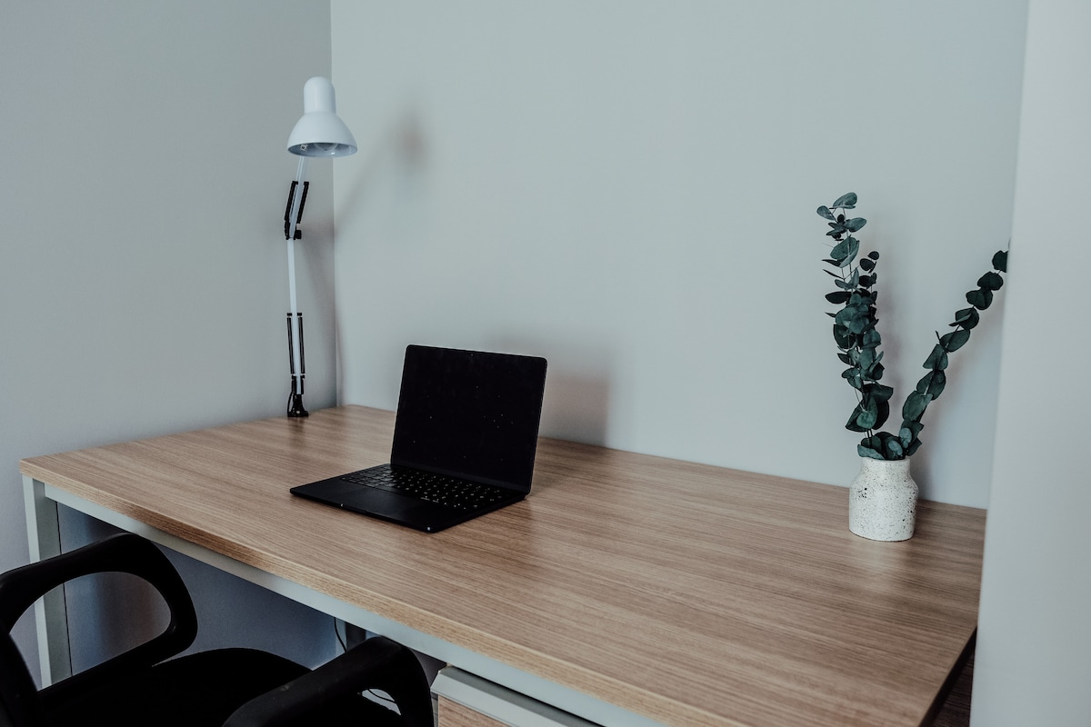 A dedicated workspace is presented with a wooden desk, a minimalist desk lamp, and a laptop. A small potted plant adds a touch of greenery, enhancing the clean and organized look. The light-colored wall creates a bright atmosphere for productivity.
