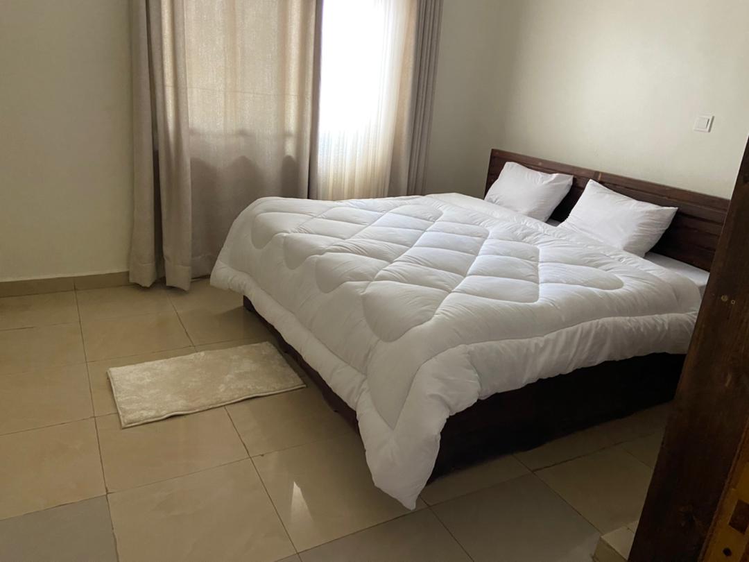A spacious sleeping area features a dark wooden bed frame with a soft, white comforter laid over the mattress. Two pillows are neatly arranged at the head of the bed. Natural light filters through a large window, complemented by light-colored curtains.