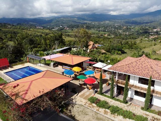 An aerial view showcases a vibrant outdoor area featuring a swimming pool surrounded by colorful umbrellas. Nearby, multiple seating areas are arranged, providing ample space for relaxation. Lush greenery and distant mountains form a scenic backdrop, enhancing the tranquil setting.