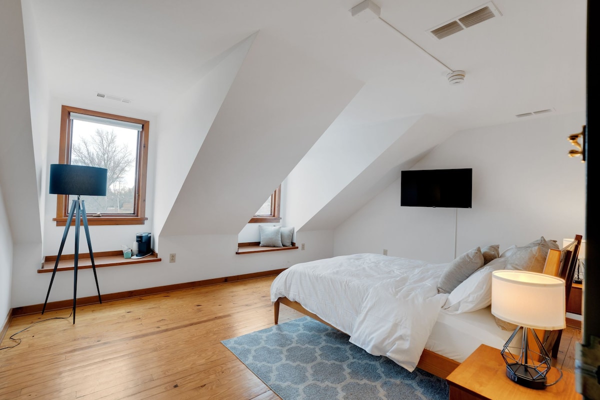 A spacious bedroom is highlighted by sloped ceilings and natural light flowing through two windows. A comfortable queen bed with white bedding is positioned next to a bedside lamp. A modern television is mounted on the wall, and a cozy area rug adds warmth to the wooden floor.